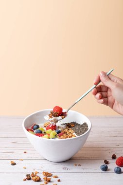 Beyaz bir kahvaltı kasesinden bir kaşık dolusu ahududu, granola ve kefir ile bir kadın tarafından kaldırıldı. Yumuşak zemin ve beyaz ahşap ile dikey kompozisyon. Boşluğu kopyala. Dengeli yemek ve günlük sağlık kavramı