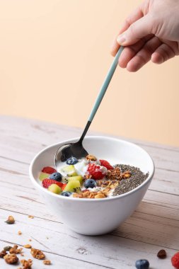 Karışık meyve, granola ve chia tohumlarıyla kaplı kefir kaplı beyaz kase, erkek eli tarafından tutulan bir kaşıkla. Pastel arka plan ve kırsal yüzey. Uzayı kopyala Günlük sağlık ve beslenme kavramı
