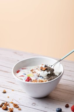 Bir kase kefir, taze meyve, granola ve chia çekirdeği içinde kaşık. Şeftali rengi arka plan ve beyaz ahşap masa. Uzayı kopyala Doğal kahvaltı ve sağlıklı yaşam tarzı kavramı.