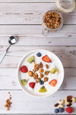 Kefir, granola, çilek, kivi, ahududu ve yaban mersinli beyaz kase bir kavanoz granola ve metal kaşığın yanında. Kırsal görünüm, dikey üst görünüm. Uzayı kopyala Sağlıklı beslenme ve modern yaşam tarzı kavramı