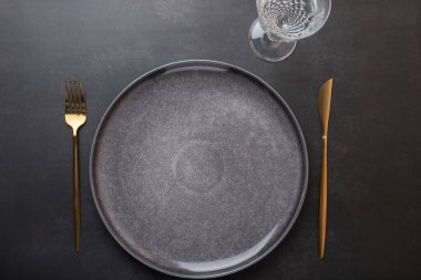 Üzerinde altın çatal bıçak ve kristal cam bulunan koyu taştan arka planda gri bir seramik tabağın üst görüntüsü. Uzayı kopyala Minimalist ve zarif bir tarz. Tablo ayarları ve çağdaş tasarım kavramı