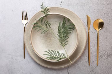 Üzerinde eğrelti otu yaprağı ve çatallı taş zemin üzerinde altın çatal bıçak bulunan bej seramik tabakaların üst görüntüsü. Doğal dekor ve zarif masa tasarımı kavramı