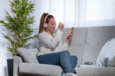 Evdeki kanepede oturan bir kadın, video görüşmesiyle kutlama yapıyor, pembe kulaklıklar ve cep telefonuyla.