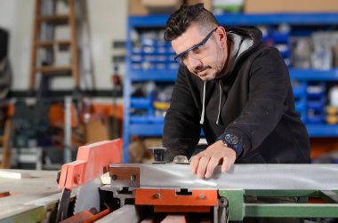 Marangoz atölyesinde testere, siyah kazak ve koruyucu gözlüklerle odun kesiyor.