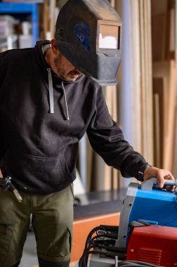 İşçi atölyede kaynak ekipmanlarını ayarlıyor, baş ve siyah iş kıyafetlerine maske takıyor