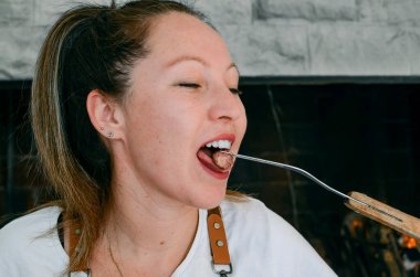 Açık gözlü, gülümseyen kadın, ön plandaki ızgaranın yanında kuzu eti yiyor.