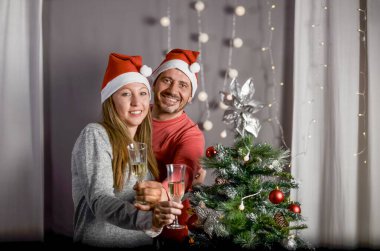 Noel 'in başlangıcını kutlayan çift, ağacın yanında, iki bardak şampanya, Noel Baba şapkaları, evde gülümsüyor.