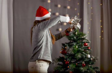 Noel ağacını kırmızı Noel Baba şapkalı ve gri arka planlı bir kadın koyuyor.