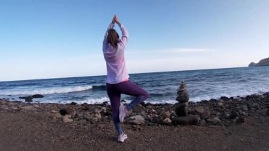 Spor kıyafetleri giyen bir kadın, arka planda dağ olan kayalık bir sahilde yoga ağacı pozu veriyor.
