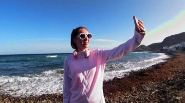 Yüzünde kulaklıklarla kayalık bir sahilde selfie çeken gülümseyen kadın. Canlı bir gökkuşağı ve okyanus dalgaları mükemmel bir yolculuk anı yaratır.