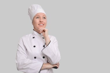 Beyaz üniformalı kadın aşçı yeni tarifler düşünüyor ve gri arka planda gülümsüyor.