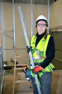 Metal çivi tutan, güvenlik kaskı ve yeleği takan inşaat işçisi kadın inşaat alanına sıva monte etmeye hazırlanıyor.