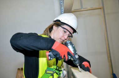 Kadın inşaat işçisi elektrikli matkap kullanıyor, inşaat sahasına alçı takıyor, güvenlik kaskı, gözlük ve eldiven takıyor.