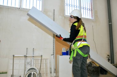 Alçıpan taşıyan kadın işçi, inşaat alanında çalışıyor, güvenlik ekipmanı takıyor.