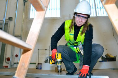 Kablosuz elektrikli matkap kullanarak alçı döşeyen kadın inşaat işçisi, güvenlik ekipmanı giyiyor.