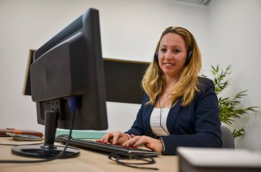 Kulaklık takan iş kadını modern ofiste masaüstü bilgisayarıyla çalışıyor.