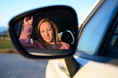 Dikiz aynası, gün batımında arabasını sürerken gülümseyen ve saçını düzelten bir iş kadınını yansıtıyor.