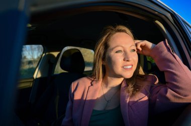 İş kadını araba camından dışarı bakıyor, yol boyunca gün batımının keyfini çıkarıyor.