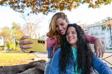 Sevgililer gününü kutlamak için parkta akıllı bir telefonla selfie çekerken sarılıp gülümseyen iki kadın.