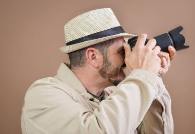 Gülümseyen fotoğrafçı bej şapka takıyor. Stüdyoda, kahverengi arka planda profesyonel kamerayla fotoğraf çekiyor.