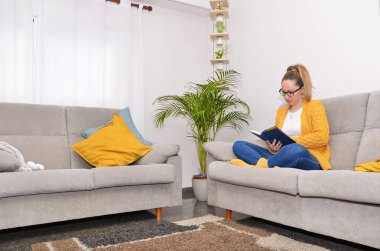 Rahat oturma odasında kitap okuyan genç bir kadın, rahat mobilyalar ve ev bitkileriyle çevrili.