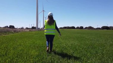 Mühendis, açık mavi bir gökyüzü altında rüzgar türbinlerinin durumunu incelemek için yeşil çimen tarlasında yürüyor.