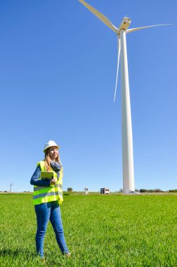 Güvenlik yeleği ve miğferi olan kadın mühendis tablet tutuyor ve mavi gökyüzü olan yeşil bir alanda rüzgar türbini denetliyor.