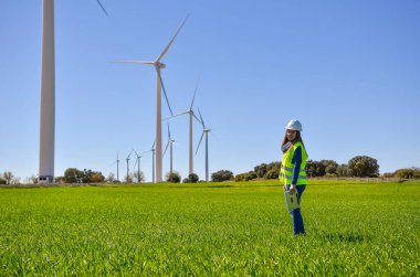 Mühendis tablet tutuyor, yeşil alandaki rüzgar türbinlerini inceliyor, yenilenebilir enerji ve sürdürülebilirlik üzerinde çalışıyor.