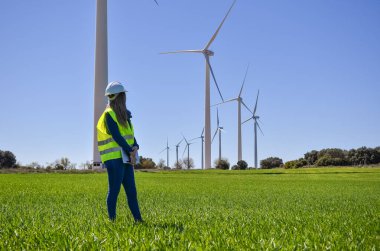 Mühendis, yenilenebilir enerji ve sürdürülebilirliği simgeleyen yeşil bir alanda rüzgar türbinlerinin yakınında çalışıyor.