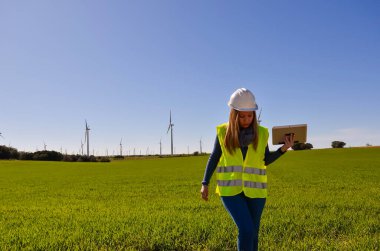 Güvenlik yeleği ve kask giyen kadın mühendis rüzgar türbini alanında tabletle yürüyor ve çalışıyor, yenilenebilir enerji kavramı