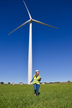Sert şapka ve güvenlik yeleği olan kadın mühendis yeşil alanda yürüyor yenilenebilir enerji ve sürdürülebilirlik için rüzgar türbinini inceliyor