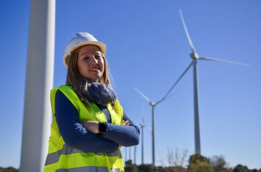 Kendine güvenen bir kadın mühendisin portresi, yenilenebilir enerji ve sürdürülebilirliği simgeleyen rüzgar türbini santralinde çapraz kollarla poz veriyor.