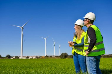 Güvenlik yeleği ve kask giyen iki mühendis, yenilenebilir enerji ve sürdürülebilirliği temsil eden yeşil bir alanda çalışan rüzgar türbinlerini gözlemliyor.