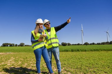 Güvenlik yelekleri ve miğferleri olan mühendisler yeşil alanda rüzgar türbinlerini analiz eden tablet kullanıyorlar, yenilenebilir enerji kavramı.