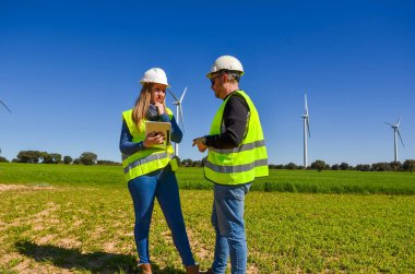 Güvenlik yeleği ve kask giyen iki mühendis yeşil alanda rüzgar türbinlerinin yanında yenilenebilir enerji ve sürdürülebilirliği destekleyen bir tablet kullanmayı tartışıyorlar.