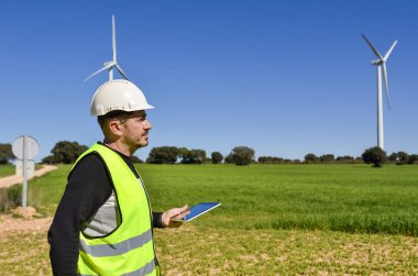 Güvenlik yeleği ve miğfer giyen mühendis tablet tutuyor ve yeşil bir alanda rüzgar türbinlerini inceliyor, sürdürülebilirlik için yenilenebilir enerji kullanıyor.