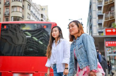 Güneşli bir günde, kırmızı bir otobüs ve binaların yakınındaki bir caddede yürüyen iki genç kadın.