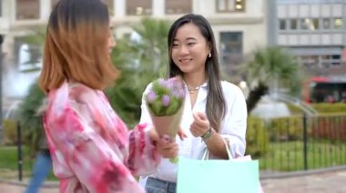 İki bayan arkadaş ellerinde çiçek buketleri ve alışveriş torbalarıyla alışveriş çılgınlığının tadını çıkarıyorlar.