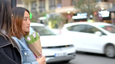 İki Asyalı turist ellerinde lale tutarken şehir caddesinde yürüyorlar.