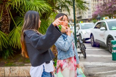 Alışveriş çılgınlığının keyfini çıkaran iki bayan arkadaş, çantaları taşıyorlar ve palmiye ağaçlarının yanındaki caddeyi işaret ediyorlar.