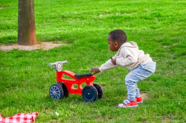 Parkta kırmızı dengeli bir bisikletin yanında çömelmiş küçük çocuk, dışarıda oyun oynamaktan zevk alıyor.