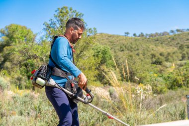 Bahçıvan, güneşli bir günde dağdaki çimleri biçmek için bir fırça makası kullanıyor.