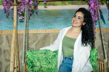 Tropikal bir ortamda kenevir yapraklarıyla süslenmiş salıncakta oturmuş gülümseyen uzun, siyah, kıvırcık saçlı genç bir kadın.