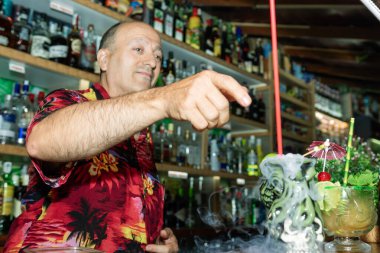 Barmen, arka planında çeşitli alkol şişeleri olan tiki barında sigara içilen Cadılar Bayramı temalı bir kokteyli işaret ediyor.