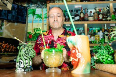 Barmen elinde tiki barında iki tropik kokteyl tutuyor, Hawaii gömleği giyiyor.