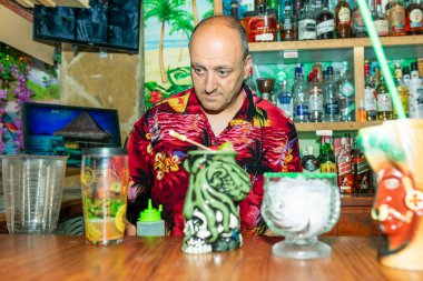 Barmen canlı bir tiki barda egzotik içecekler hazırlıyor. Etrafı renkli süslemeler ve tam bir içki sergisi ile çevrili.