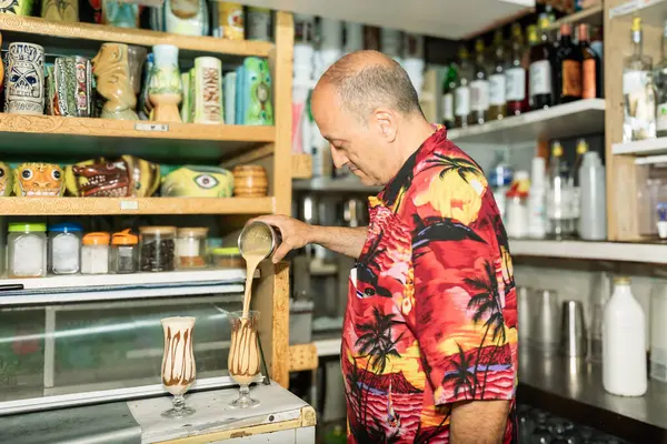 Barmen tiki bardakları ve şişelerle çevrili bir tiki barında kasırga bardaklarına kremalı kokteyl dolduruyor.