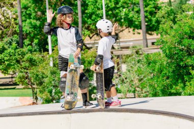 Kasklı ve koruyucu teçhizatlı iki çocuk kaykaylarını bir kaykay parkının tepesinde sallıyorlar.