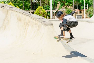 Kaykay parkında kaykay antrenmanı yapan, koruyucu kıyafetler giyen ve numaralar yapan bir çocuk.