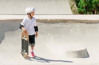 Emniyet teçhizatı takan kaykaycı çocuk kaykayını beton bir kaykay kasesinin kenarında tutuyor.